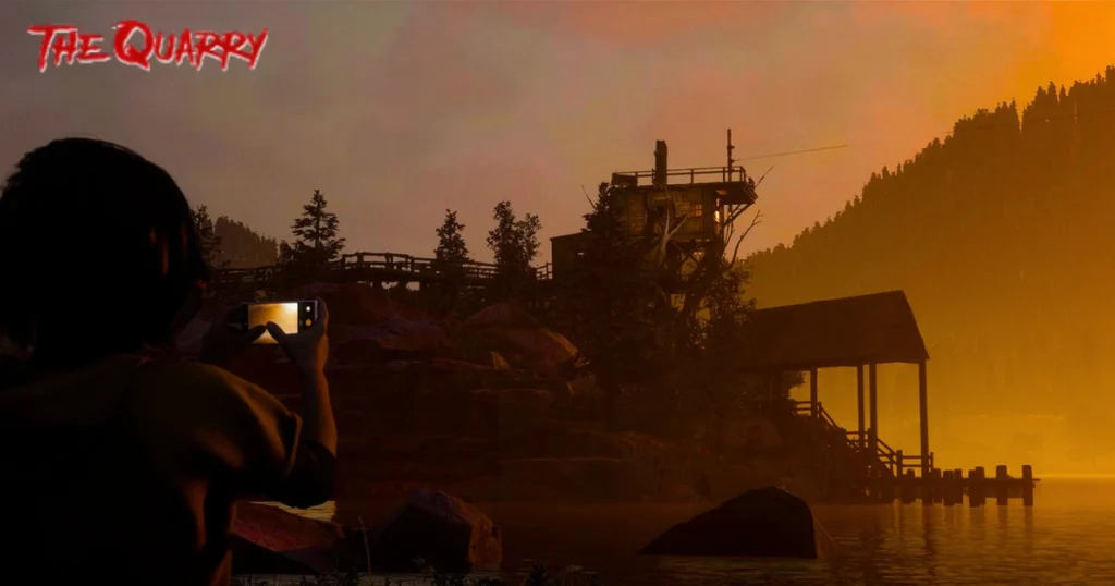 A video game screenshot from The Quarry, depicting a young woman taking a photo of a secluded cabin on a lakeshore. The setting sun casts a warm, ominous glow, creating a sense of impending danger. The cabin, perched on a cliff, appears isolated and mysterious.